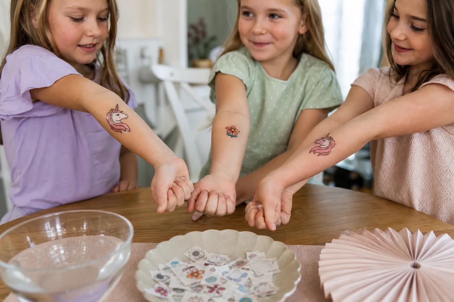 En vuxen trycker en fuktad pappersservett mot ett tatueringsmotiv på ett barns underarm vid ett dukat kalasbord, med uppklippta motiv i en skål bredvid.