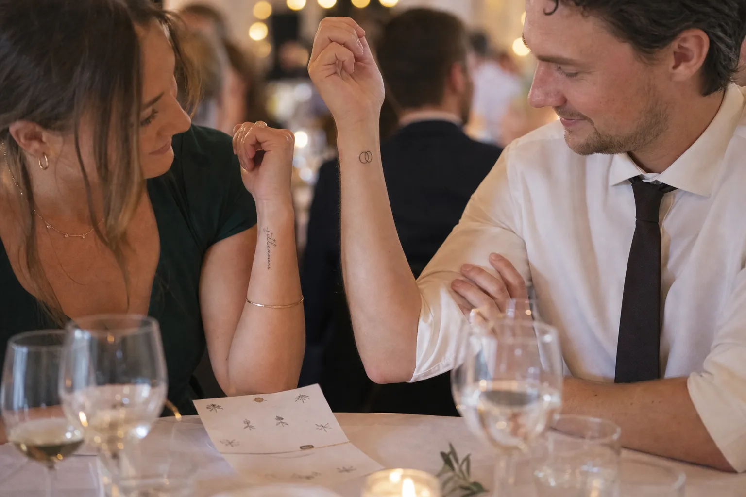 Två bröllopsgäster vid ett dukat bord håller upp underarmarna och jämför varsitt litet tillfälligt tatueringsmotiv, med ett uppklippt ark på bordet mellan dem.
