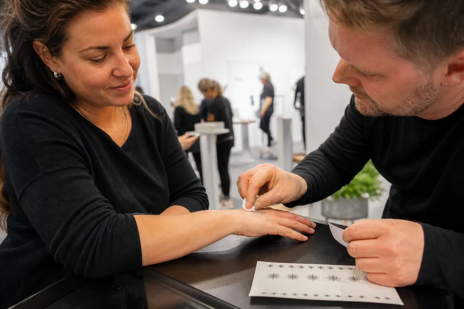 En montervärd trycker en fuktad servett mot ett litet tatueringsmotiv på en mässbesökares handrygg, med ett uppklippt ark i olika motivbredder på disken.