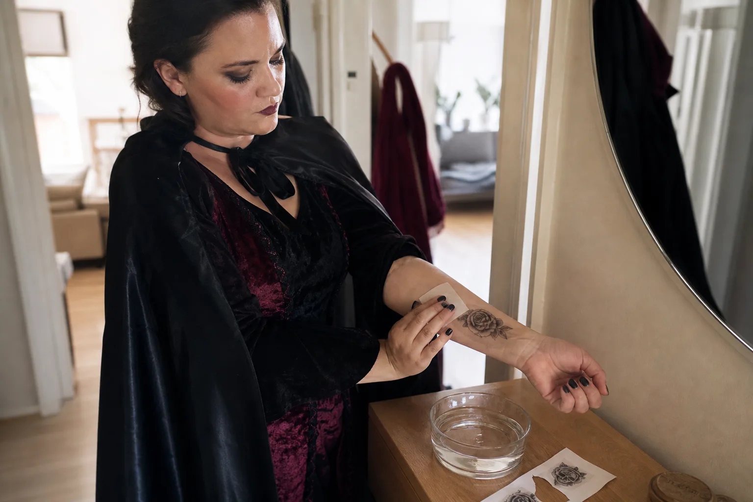 En vuxen i halloweenkostym trycker en fuktad servett mot ett tillfälligt tatueringsmotiv på underarmen framför en spegel, med ett uppklippt ark på byrån bredvid.
