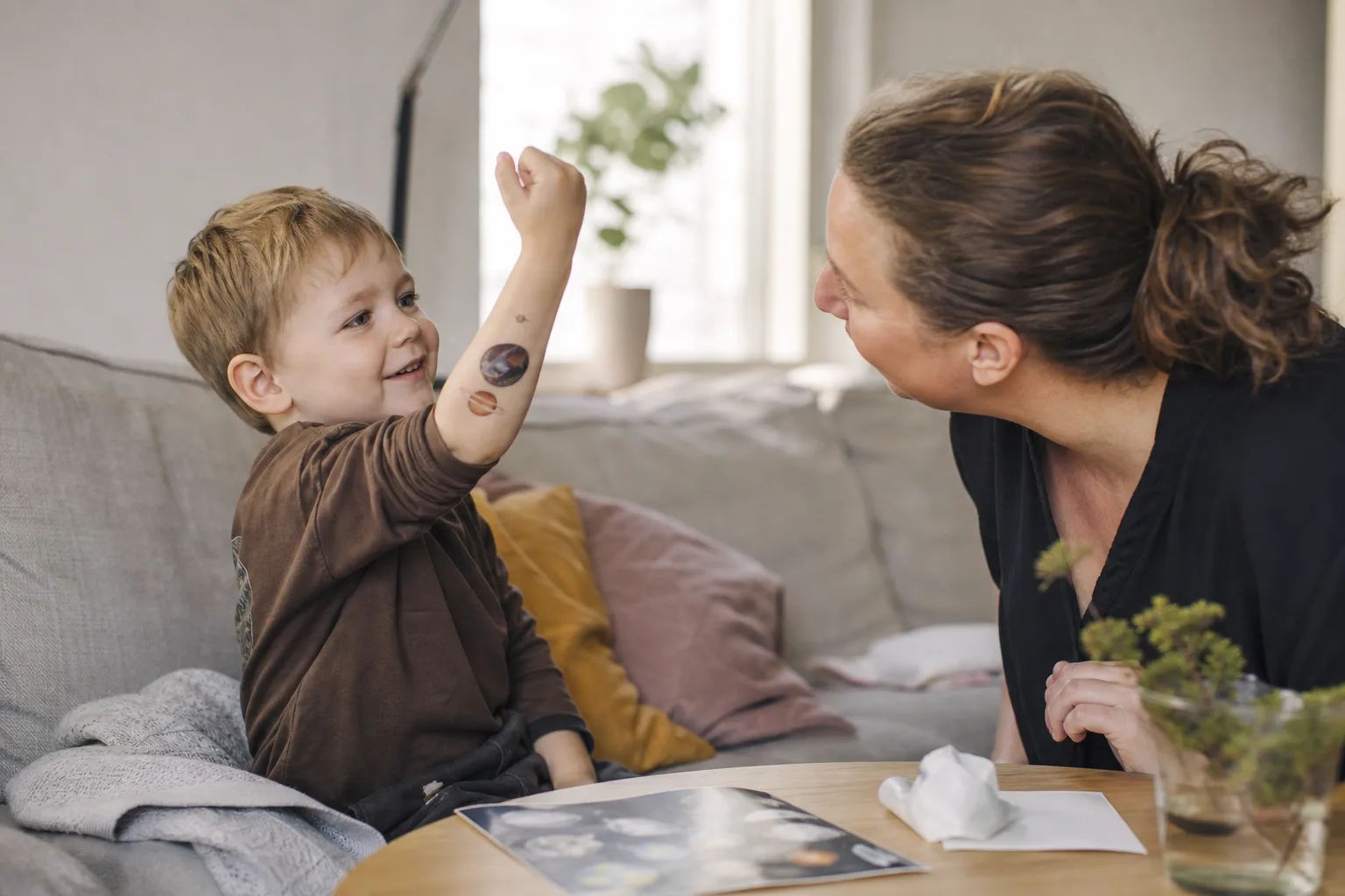 Ett barn sitter i en soffa och håller upp underarmen med ett nygnuggat rymdmotiv mot en förälder, med arket och en fuktad tvättlapp på soffbordet.
