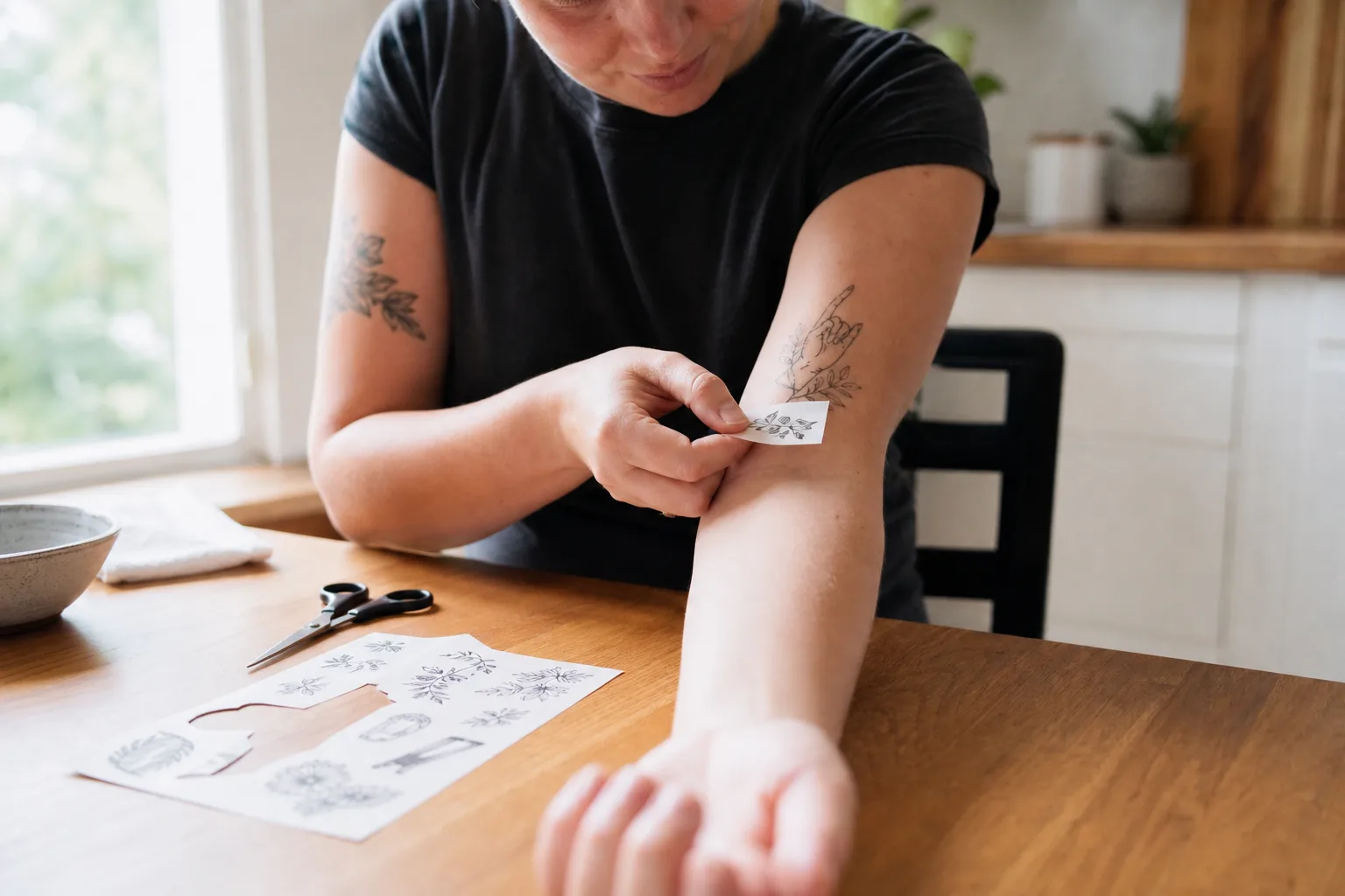 En person trycker en fuktad servett mot ett tillfälligt tatueringsmotiv på underarmen, med ett uppklippt ark på köksbordet bredvid.